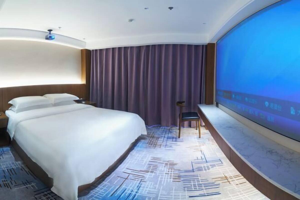 Ein Hotelzimmer mit Bett, Schreibtisch, Stuhl, großem Bildschirm und Vorhang.
