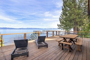 Una terraza de madera con sillas de mimbre y mesas de picnic, con vistas a un cuerpo de agua y montañas.