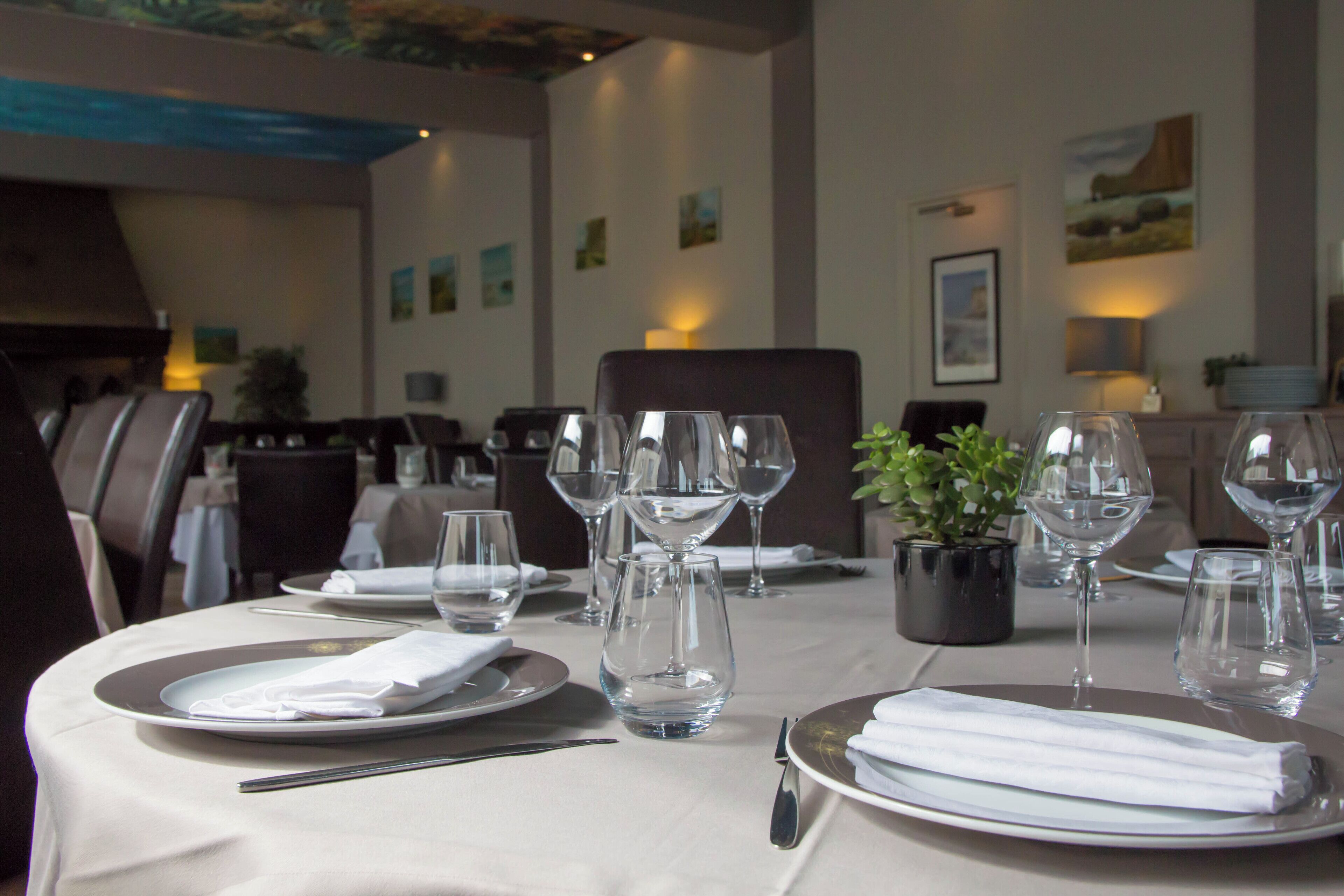 A dining table set with plates, cutlery, and wine glasses, with a potted plant and artwork on the walls.