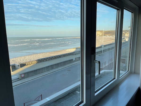 Une vue depuis une fenêtre donnant sur une plage, avec une route et une barrière.