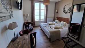 A hotel room with a bed, a desk, a chair, a lamp, a mirror, and a view of the sea through the window. - Les Fregates (Veulettes-sur-Mer)