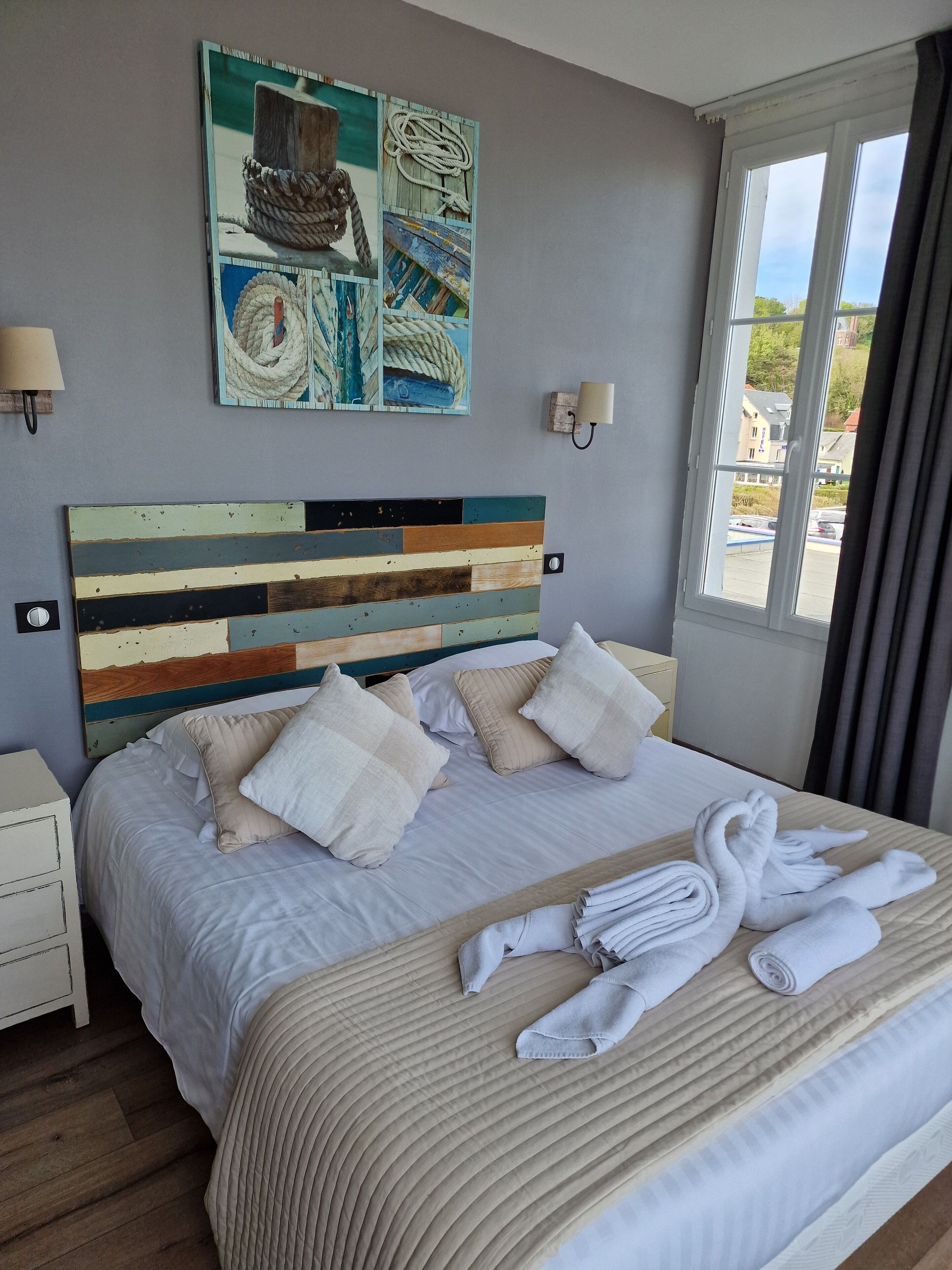 Een net opgemaakt bed met kussens en opgerolde handdoeken, een houten hoofdbord met streepdessin, een aan de muur hangend kunstwerk en een raam met gordijnen.
