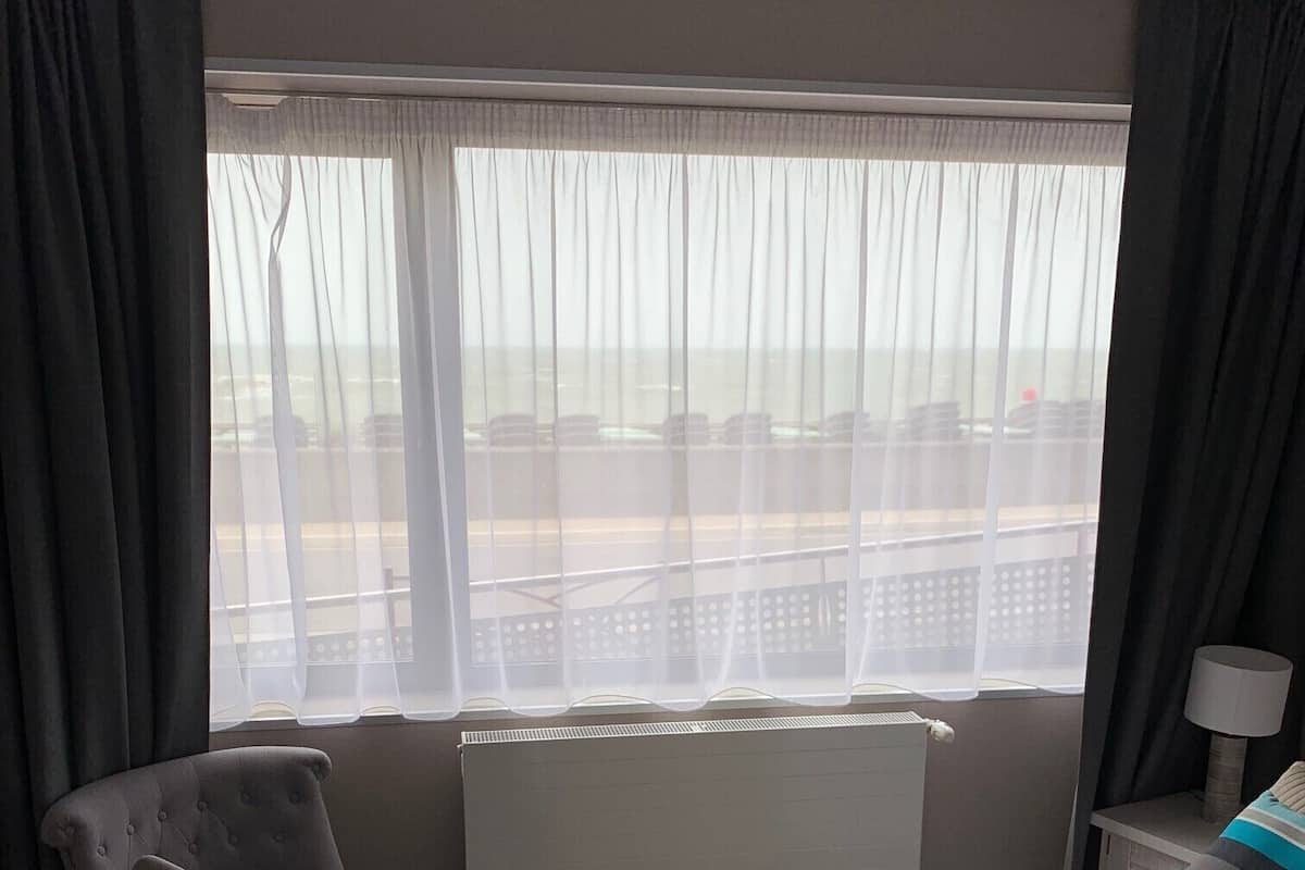 Une chambre d’hôtel avec un lit, une chaise, une fenêtre avec des rideaux, un radiateur et une lampe.