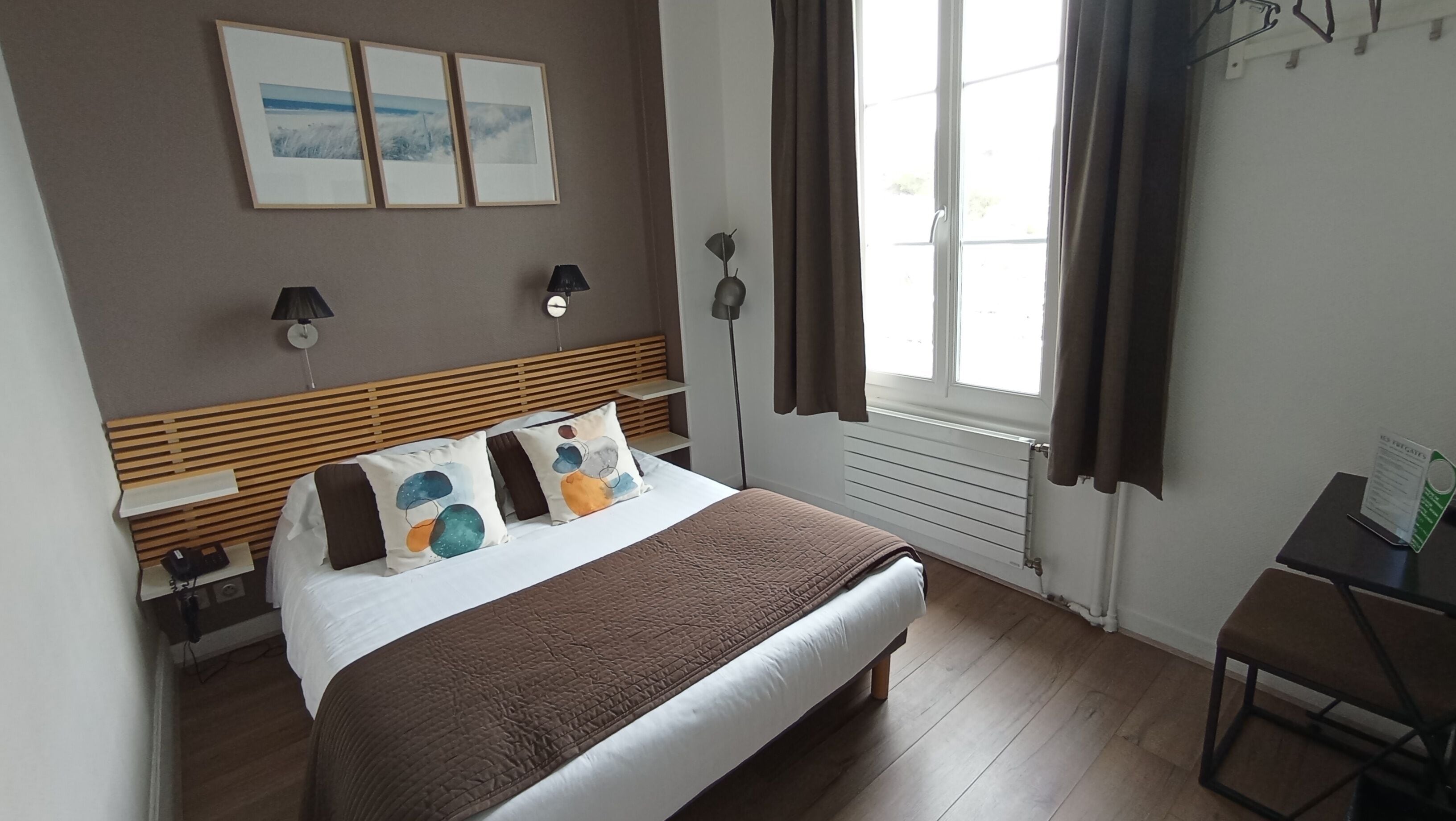 A bedroom with a bed, bedside tables, a lamp, a window with curtains, and a wall with framed pictures.