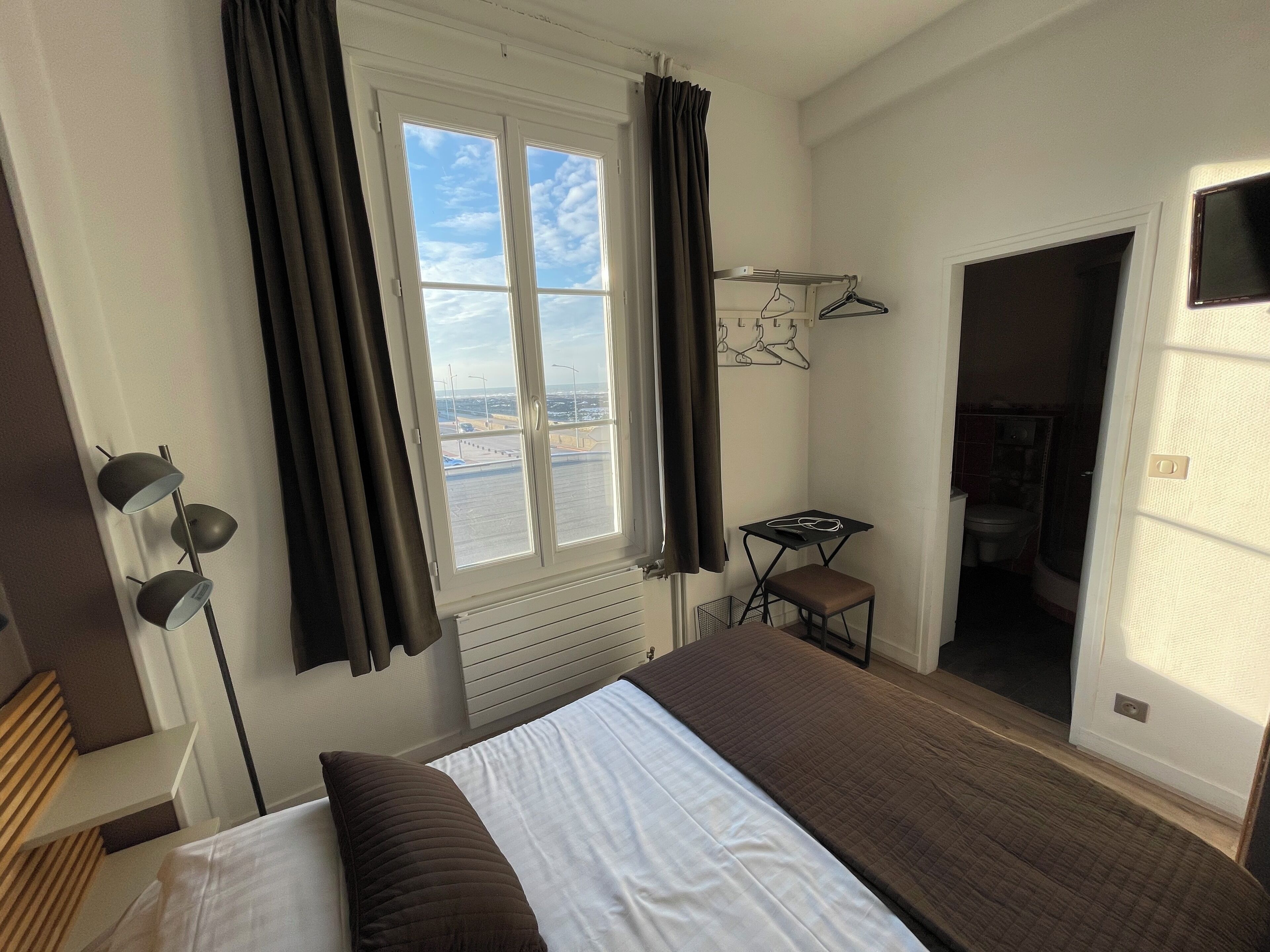 Habitación de hotel con cama, una mesita, una silla, perchero y vistas al exterior.