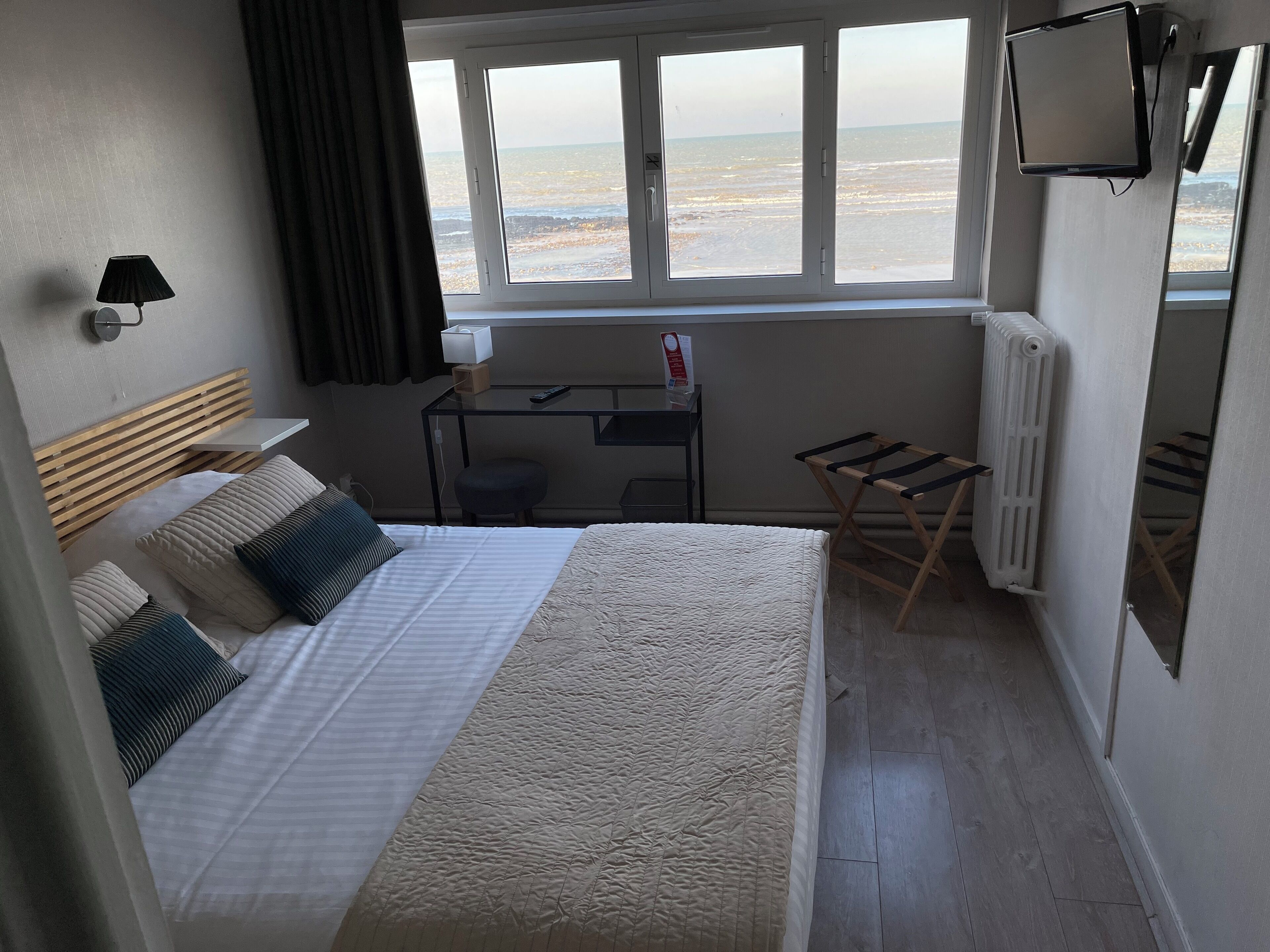 A bedroom with a bed, a desk, a chair, a radiator, and a window with a view of the beach.