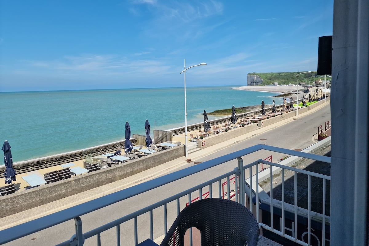 Une vue sur la mer depuis un balcon donnant sur une plage avec des transats et des parasols, une promenade et une falaise au loin.