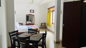 A hotel room with a bed, a desk, and a chair. There is a window with curtains, a door, and a mirror on the wall. - Sharon Inn (Kandy)