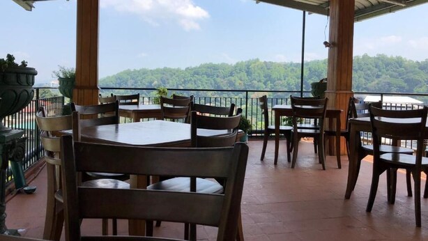 An outdoor terrace with wooden tables and chairs, a metal railing, and a view of a forested hill. - Sharon Inn (Kandy)