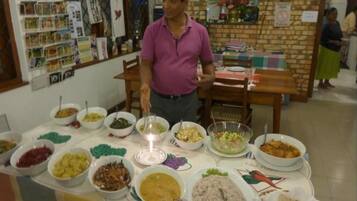 Una mesa de buffet con diversos platos, un hombre de pie al frente, y un muro de ladrillo con una ventana y adornos al fondo.
