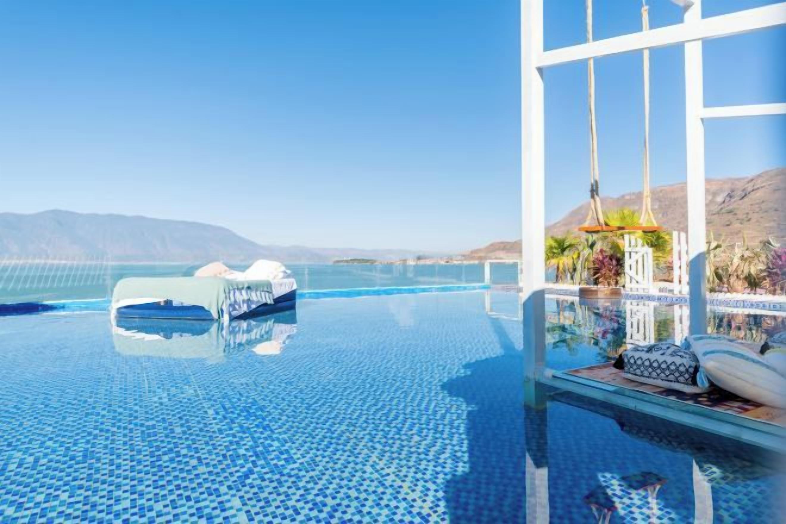 Una piscina infinita con agua azul cristalina, un área de descanso con barandilla blanca y vistas a montañas distantes.