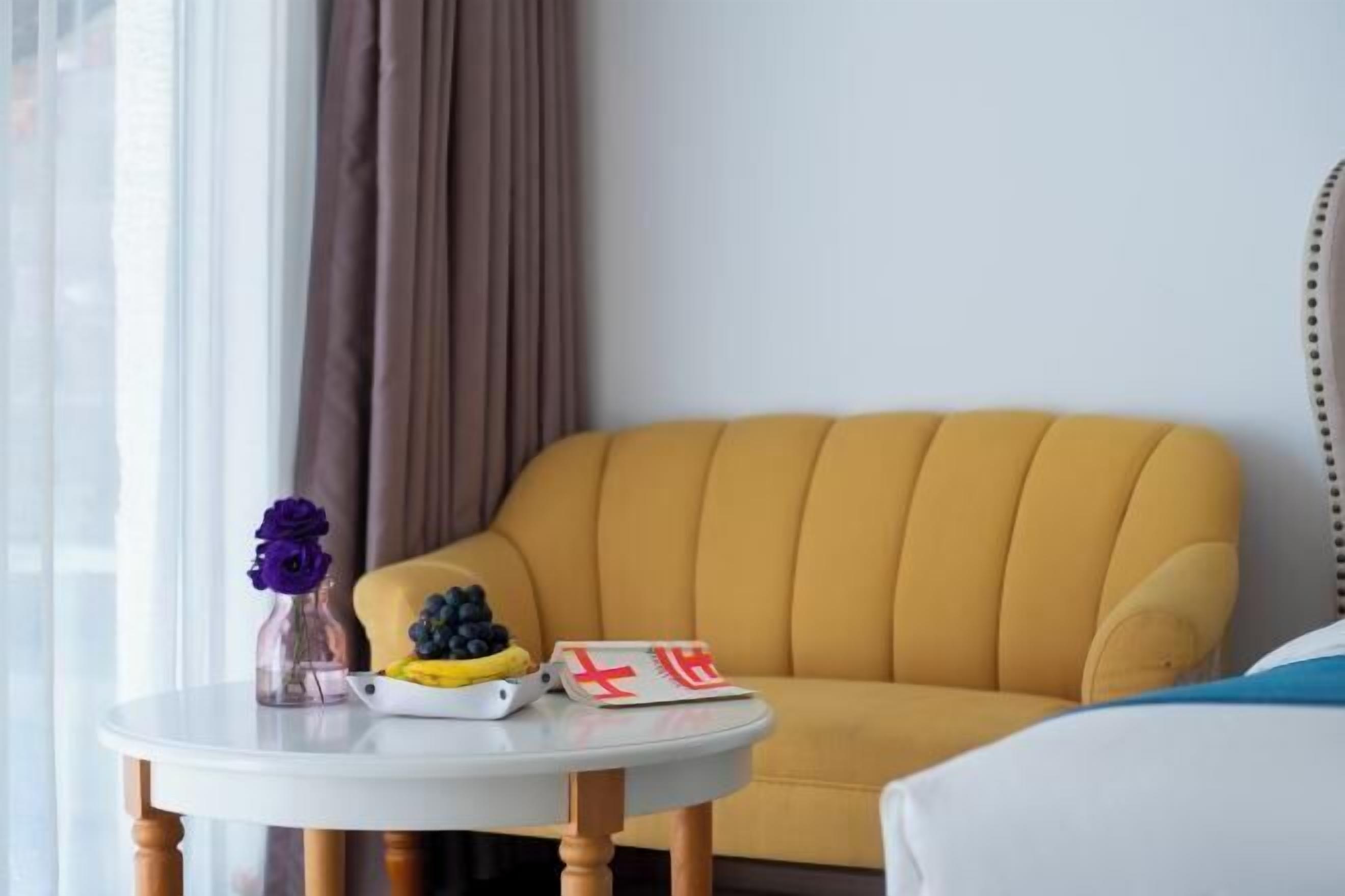 A yellow armchair next to a white table with a vase of purple flowers, a bowl of fruit, and a magazine.