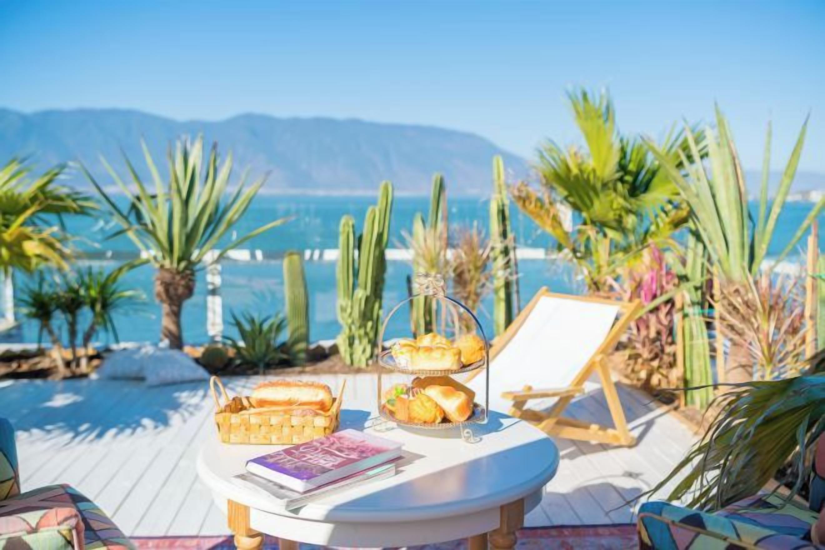 A sunlit outdoor seating area with a view of the sea and mountains, featuring a table with a book and a basket of bread.