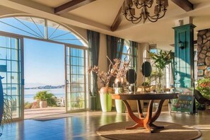 Una habitación amplia con un gran ventanal que da a una vista panorámica, una mesa redonda con un jarrón de flores y un mueble de color turquesa.