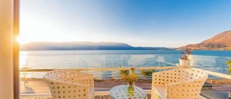 A balcony with wicker chairs and a table, overlooking a calm body of water and distant mountains.