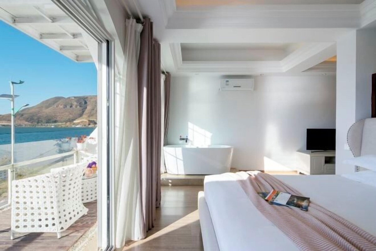 Habitación de hotel moderna con una cama grande, una bañera, un televisor y un balcón con vistas al mar.