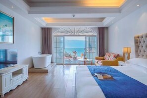 Habitación de hotel con cama, televisión, bañera, zona de estar y vista al mar, además de un ventanal grande.