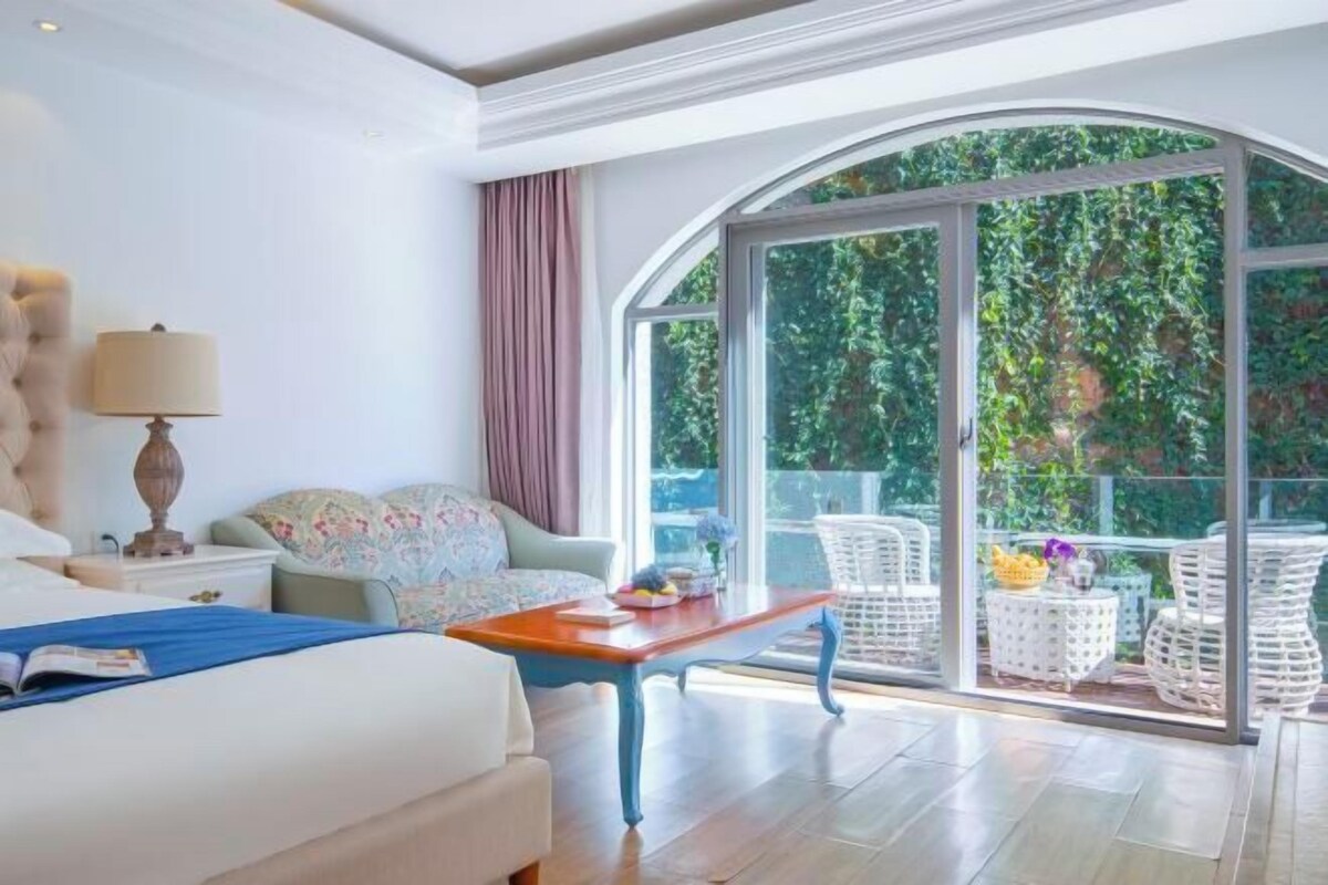 Habitación de hotel con un ventanal grande, una cama, una mesita de noche con lámpara y un pequeño balcón con mobiliario de mimbre.
