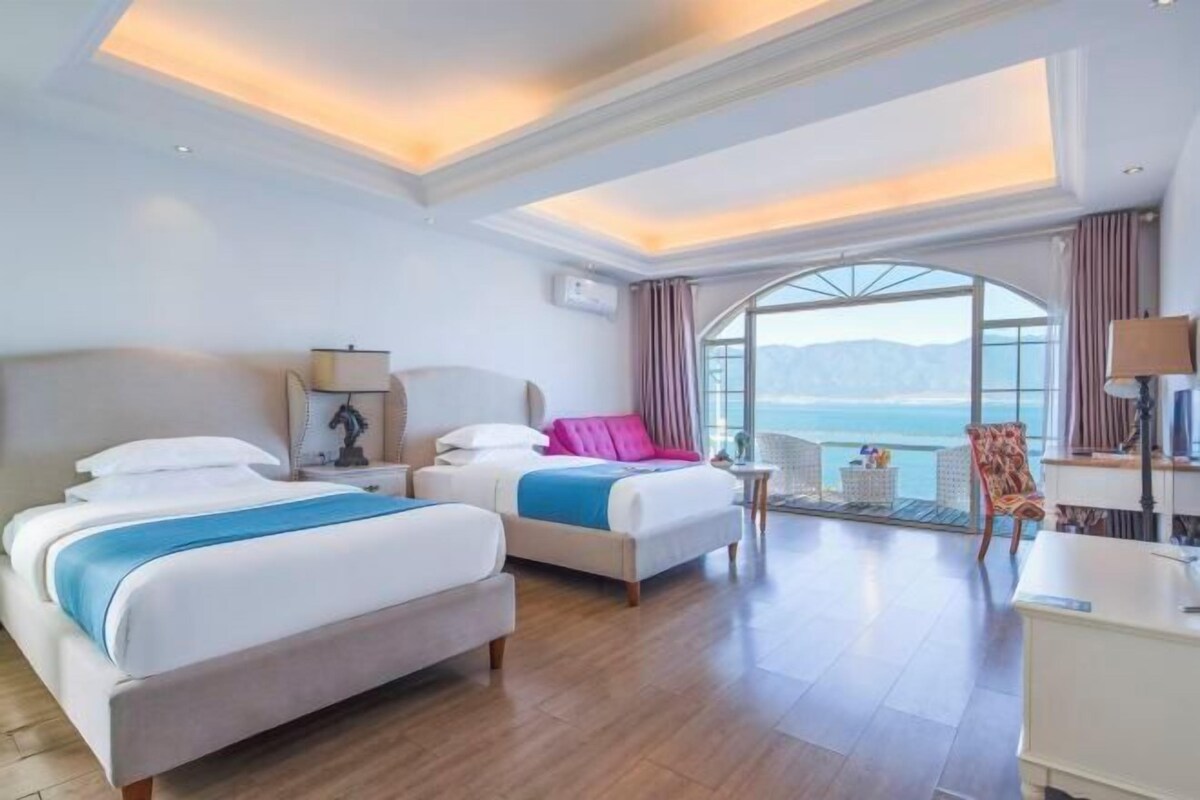 Habitación de hotel con dos camas, un escritorio, una silla y un ventanal con vista al mar.