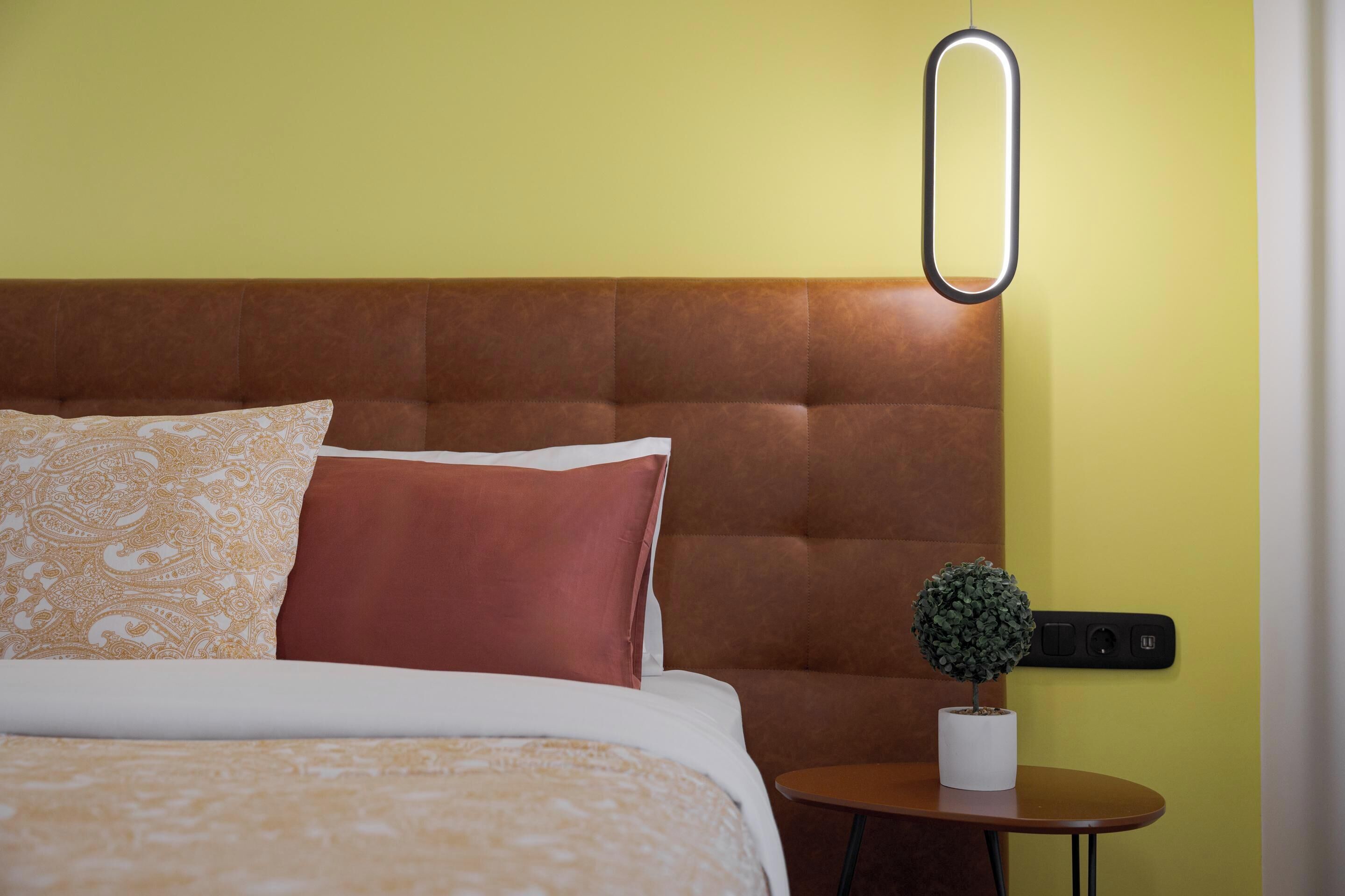 A hotel room with a tufted headboard, a bed with patterned bedding, a small table with a potted plant, and a wall-mounted light fixture.