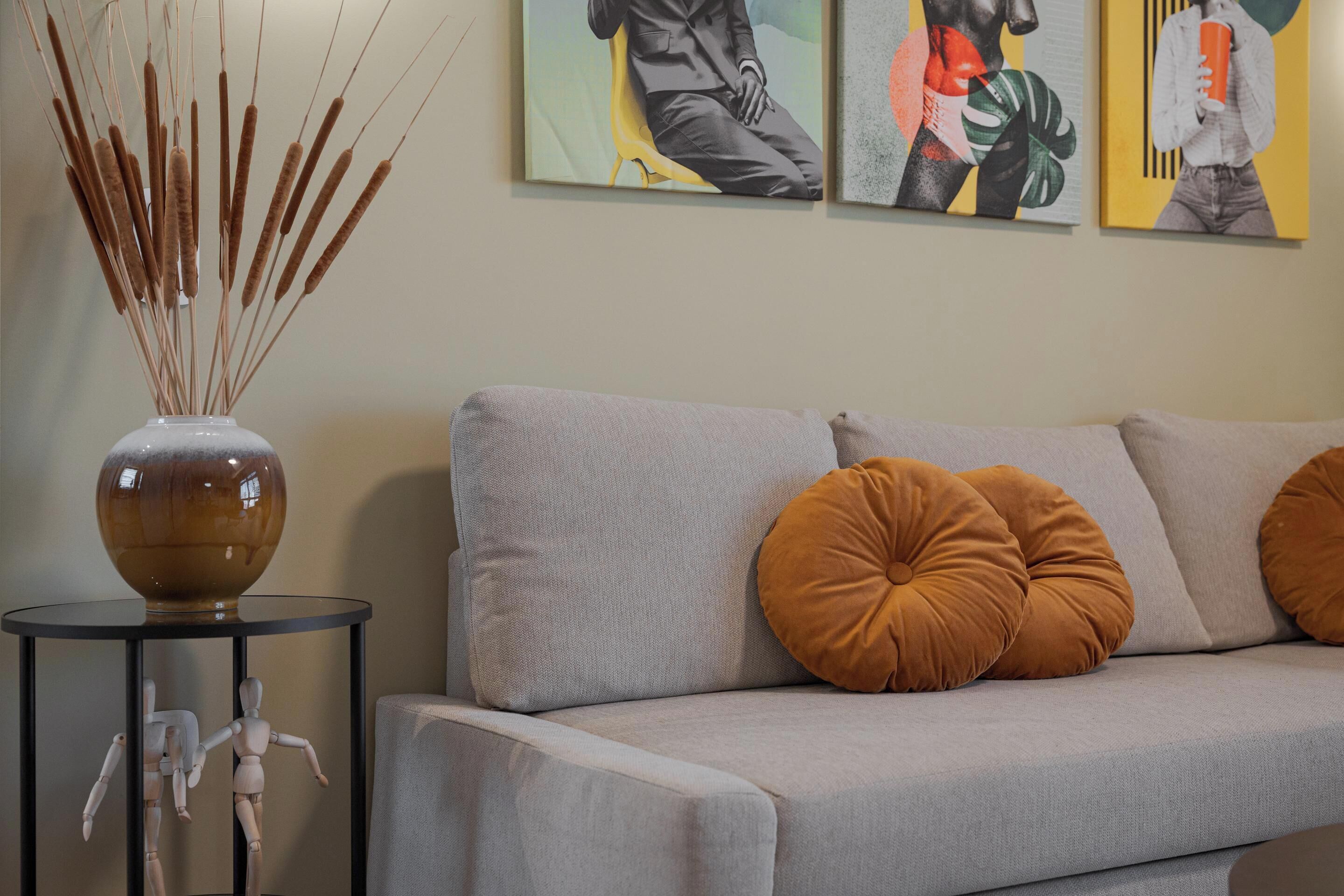 Ein modernes Wohnzimmer mit einem grauen Sofa, zwei senfgelben Kissen, einer Vase mit Schilf und drei abstrakten Bildern an der Wand.