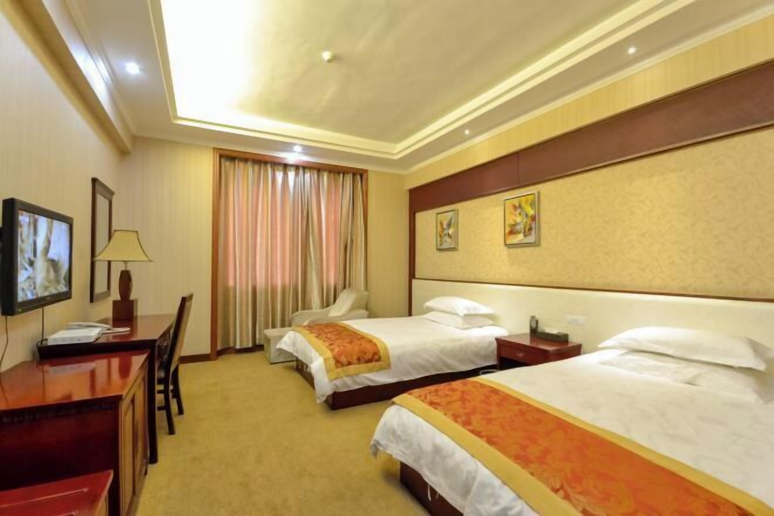 A hotel room with two beds, a desk, a TV, and a window with curtains.