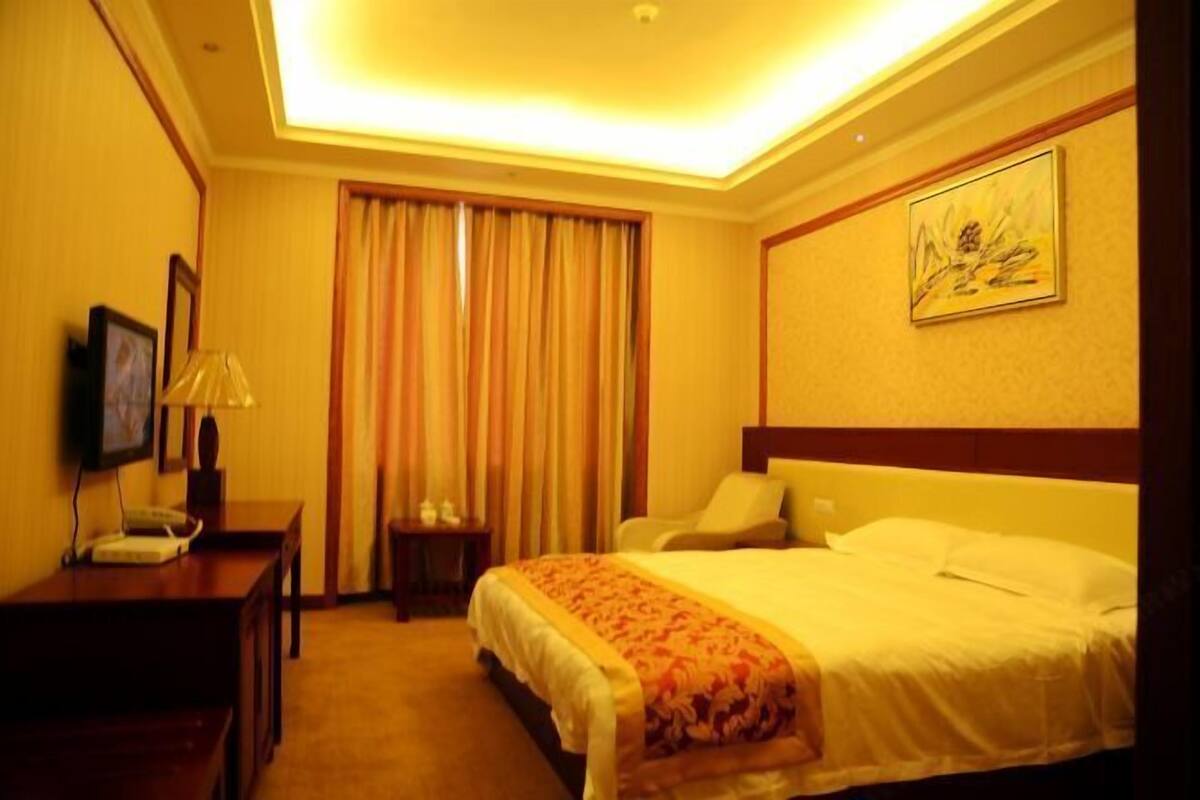 A hotel room with a bed, a desk with a lamp, a chair, and a painting on the wall.