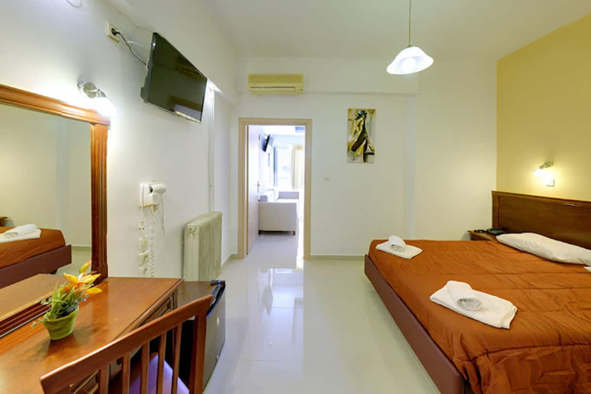 A hotel room with a wooden desk, a bed with an orange bedspread, a TV mounted on the wall, and a hallway leading to another room.