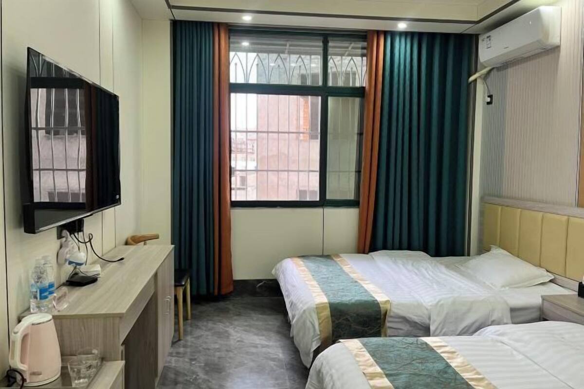 Ein Hotelzimmer mit zwei Betten, einem Fernseher, einem Schreibtisch mit Wasserkocher und einem Fenster mit Vorhängen.