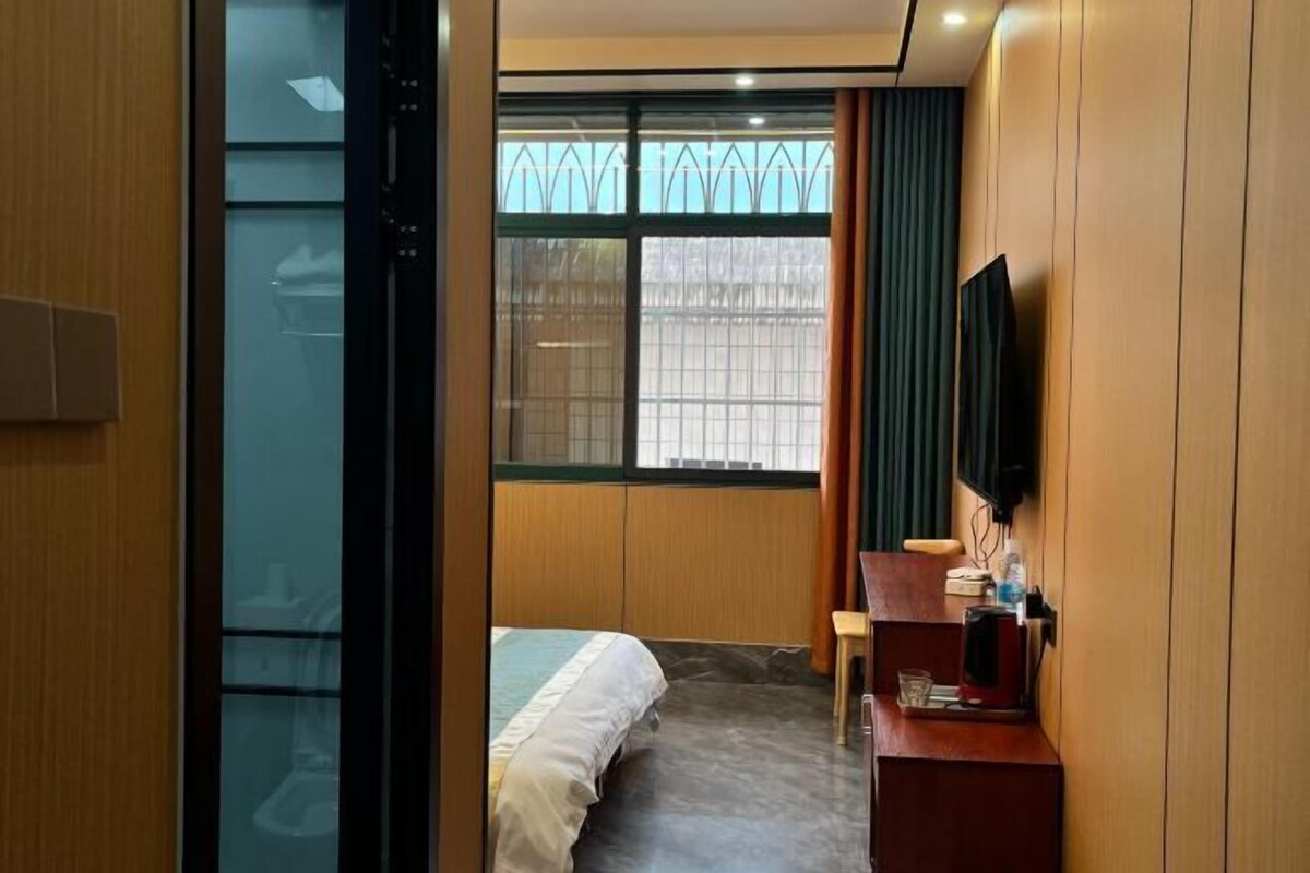 A modern hotel room with a bed, a desk with a coffee maker, a TV, and a window with blinds.