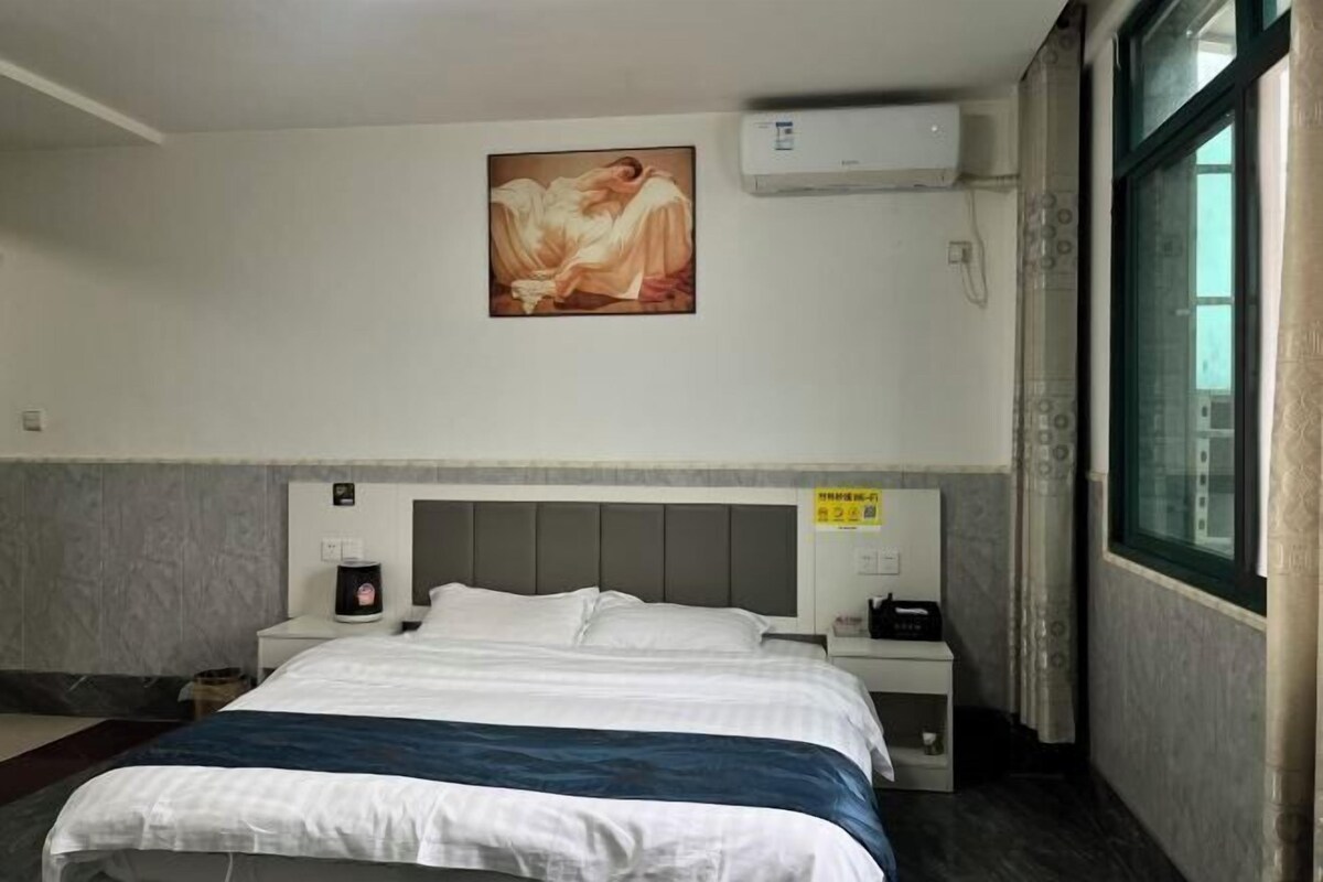 A hotel room with a bed, a painting on the wall, an air conditioner, a nightstand, and a window.
