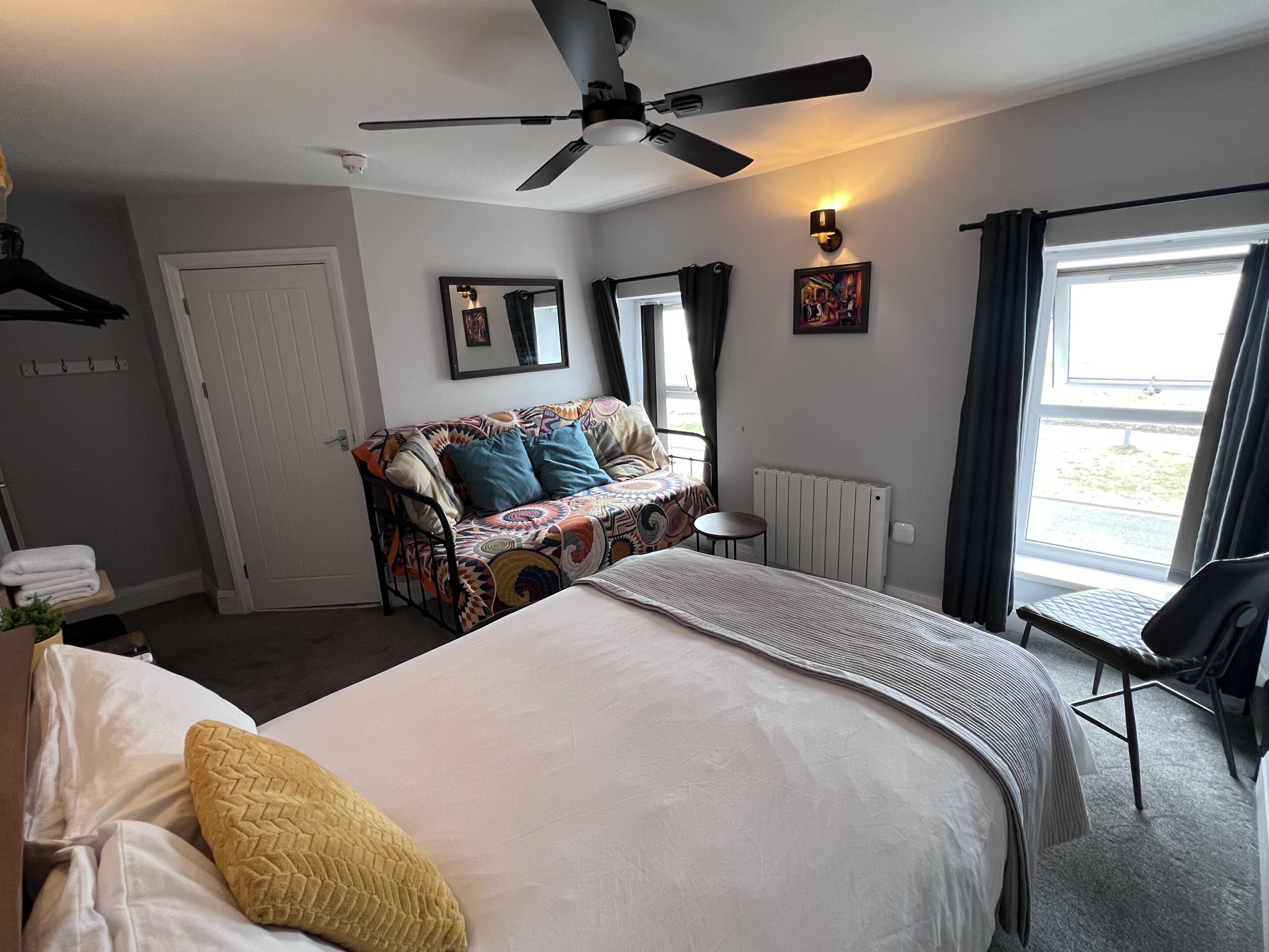 Une chambre d’hôtel comprenant un lit, un canapé, une petite table, une chaise, un ventilateur de plafond, une fenêtre avec des rideaux et un radiateur.