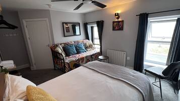 Une chambre d’hôtel comprenant un lit, un canapé, une petite table, une chaise, un ventilateur de plafond, une fenêtre avec des rideaux et un radiateur.