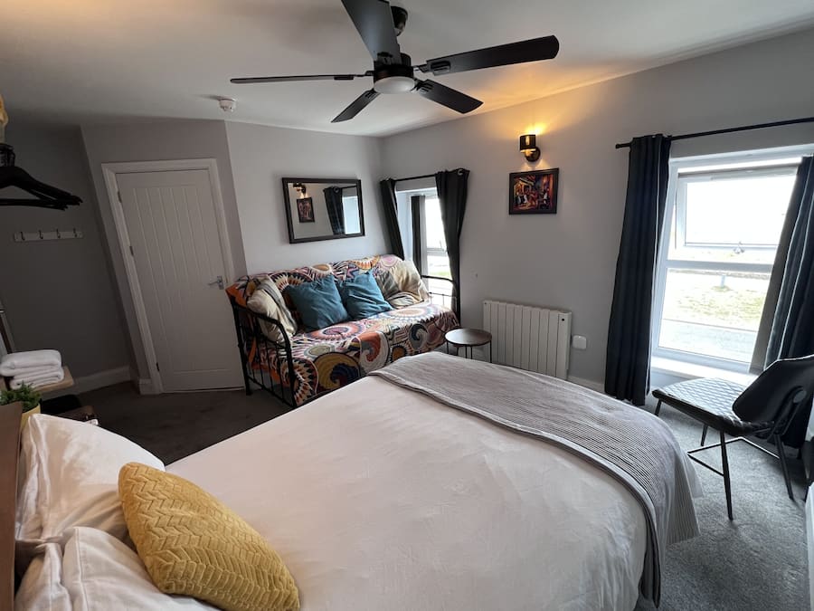 A hotel room with a bed, a sofa, a small table, a chair, a ceiling fan, a window with curtains, and a radiator.