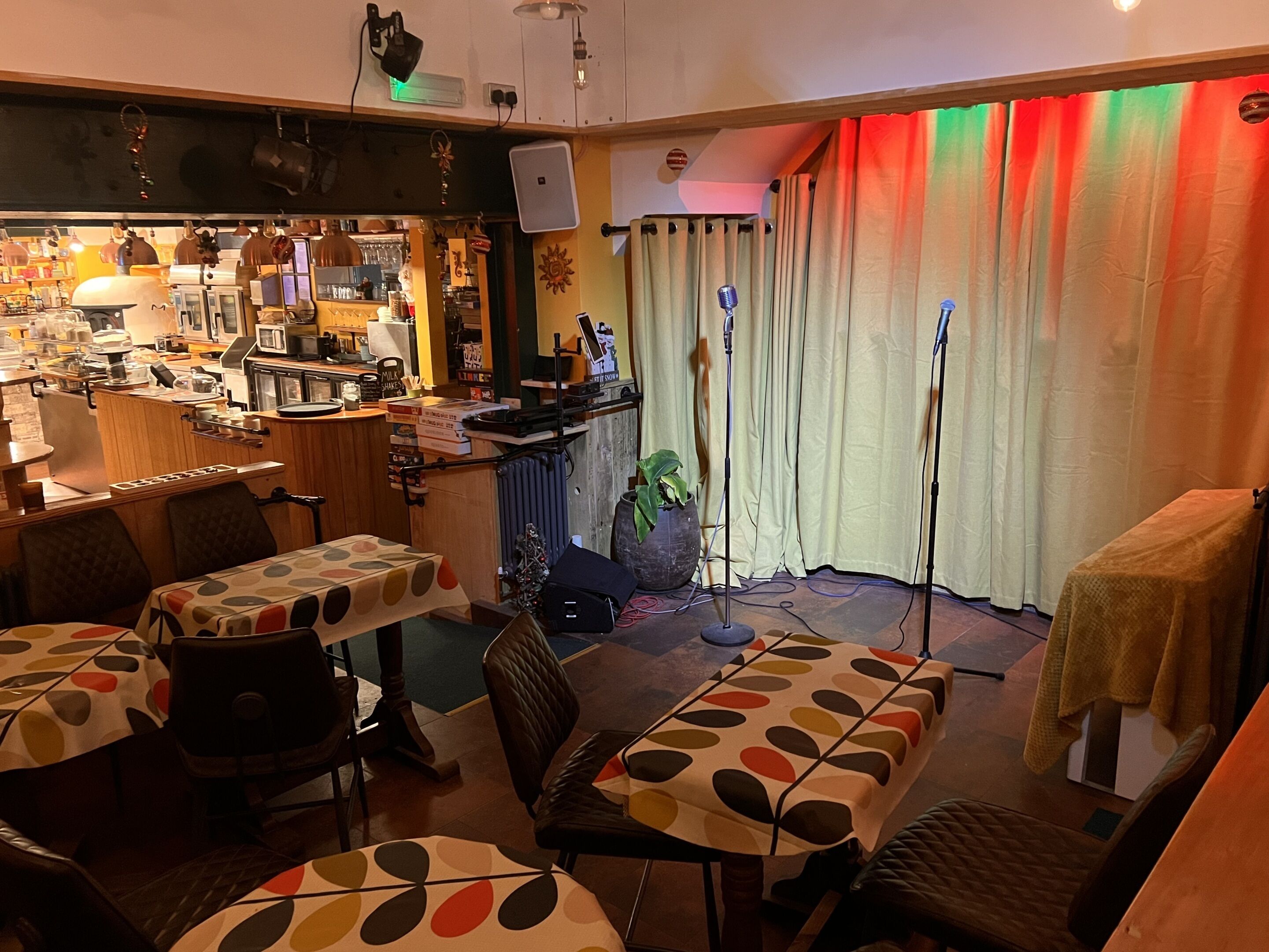 Un restaurante acogedor con zona de bar, mesas con manteles estampados y un escenario con micrófonos.