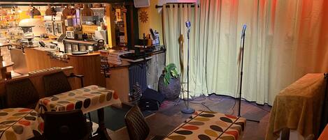 Un restaurante acogedor con zona de bar, mesas con manteles estampados y un escenario con micrófonos.