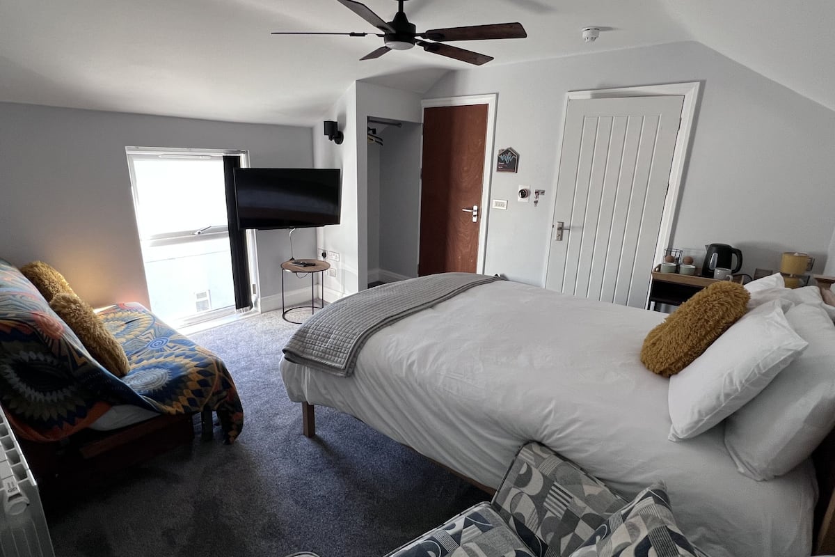 Une chambre à coucher avec un lit, un ventilateur de plafond, une télévision, une porte et une petite table sur laquelle se trouve une bouilloire.
