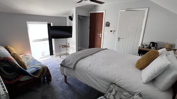 A bedroom with a bed, a ceiling fan, a TV, a door, and a small table with a kettle.