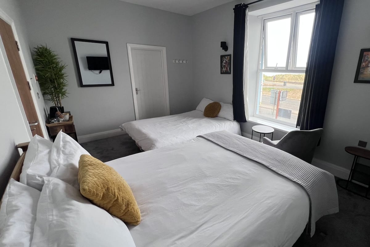 Une chambre d’hôtel avec deux lits, une petite table, une chaise, une fenêtre avec des rideaux, un radiateur et une plante en pot.