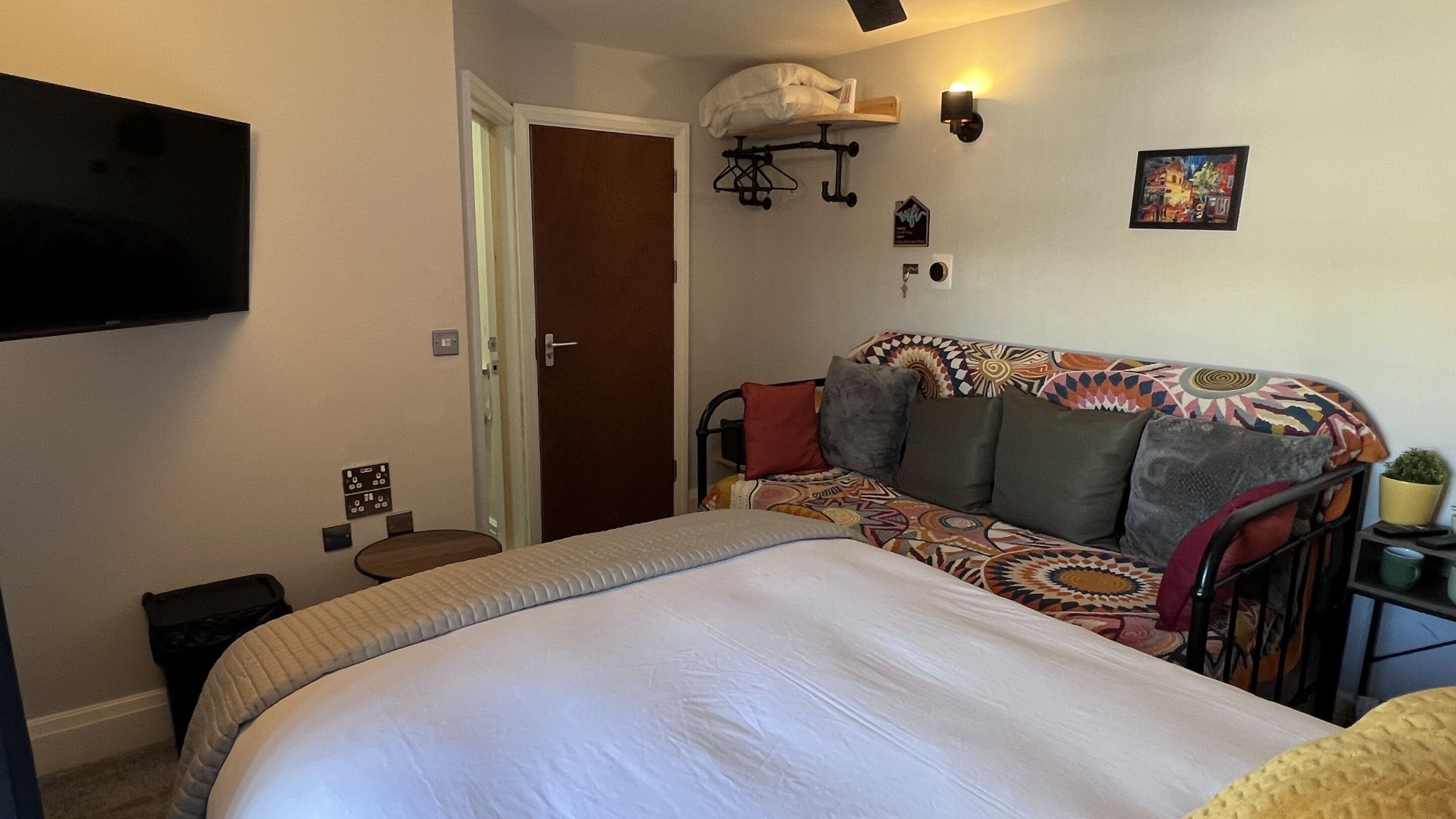 Ein Schlafzimmer mit Bett, einer Couch, einem Fernseher, einer Tür und einem an der Wand befestigten Regal mit einer Pflanze.