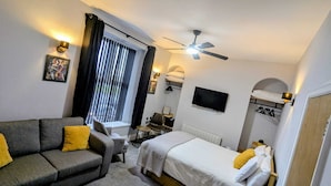 Een kleine hotelkamer met een bed, een slaapbank, een bureau, een stoel, een televisie en een plafondventilator.