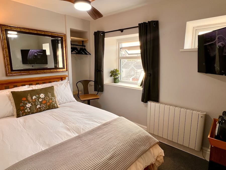 Habitación de hotel con cama, televisor montado en la pared, ventana con cortinas, radiador y una pequeña mesa con una tetera.