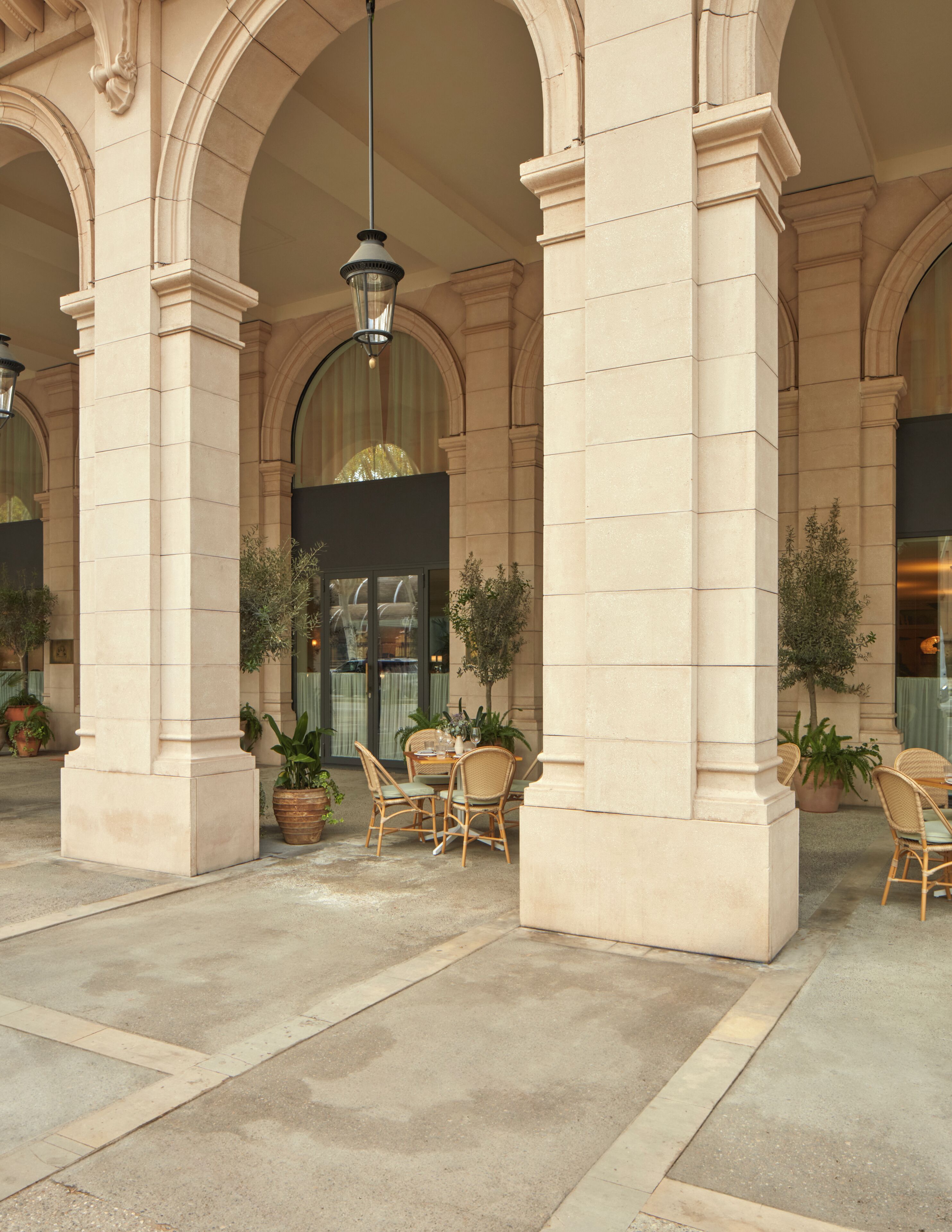 Amplio espacio al aire libre con arquitectura clásica, que cuenta con grandes columnas, una zona de estar central con sillas y una mesa, y plantas en macetas.