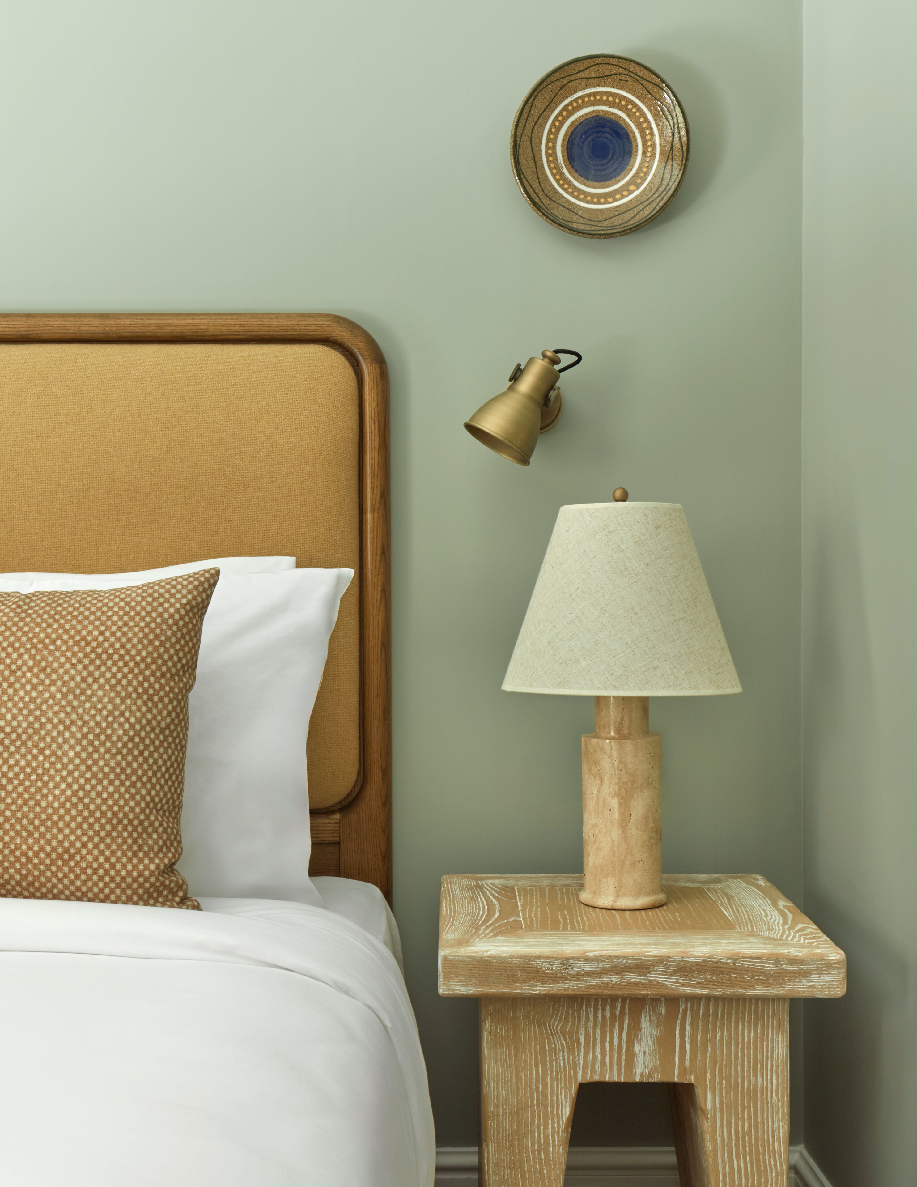 A neatly made bed with a wooden headboard, a small wooden nightstand with a lamp, and a decorative plate on the wall.