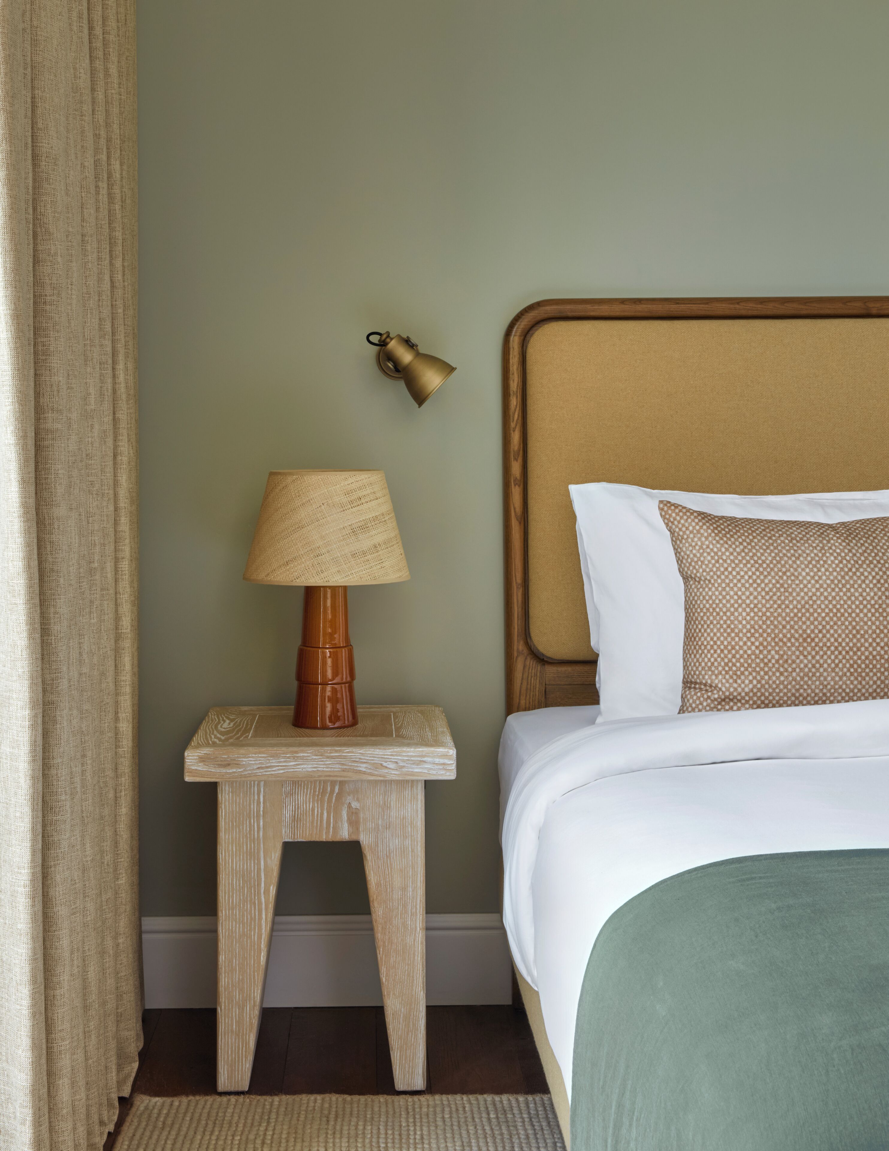 A neatly made bed with white linens and a green throw, a wooden nightstand with a lamp, and a wall-mounted light fixture.