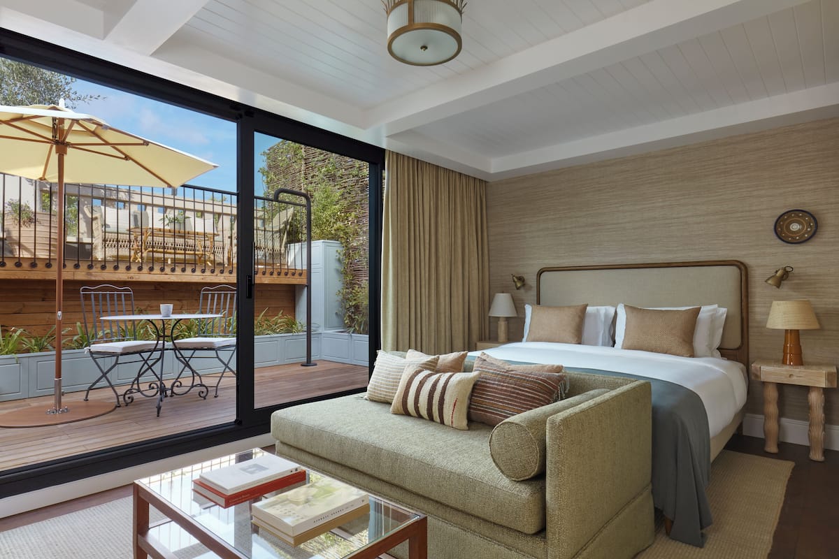 A modern hotel room with a large bed, a sofa, a dining table, and a view of the outdoor deck.