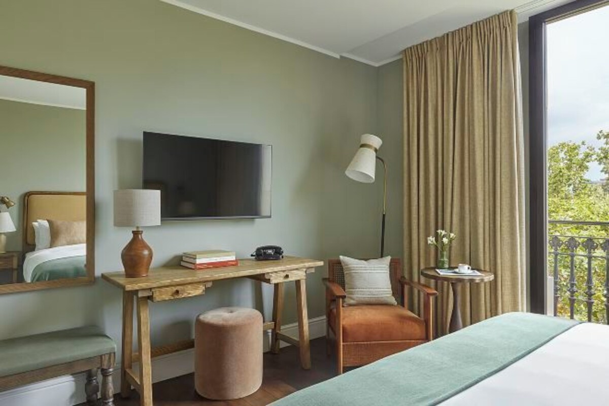 A hotel room with a bed, a desk, a chair, a small table, a lamp, and a TV.