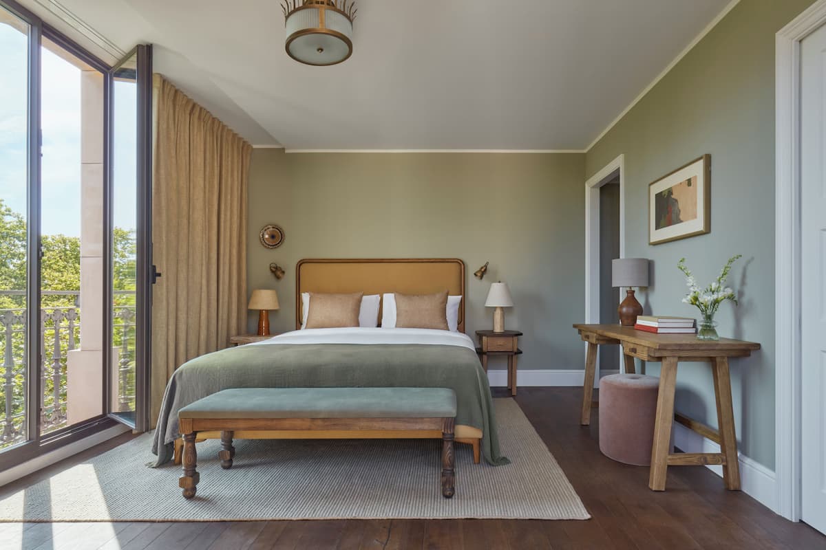 A bedroom with a large bed, a bench, a bedside table with lamps, a small stool, and a view of the outdoors.
