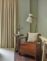 Quarto com uma cadeira de madeira, uma mesa pequena com um vaso de flores, uma luminária e uma cortina.