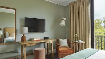 A hotel room with a bed, a desk with a lamp, a chair, a small table with a vase, and a large window with curtains.